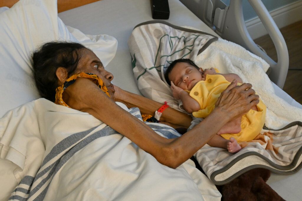 A newborn baby in a yellow romper lays sleeping beside his grandmother.